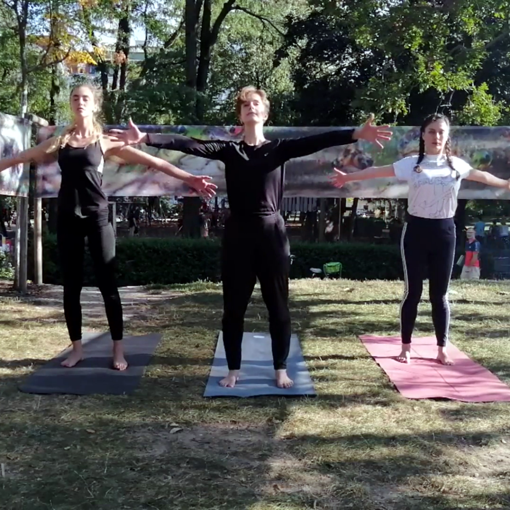 Three female dancers stand on yoga mats facing the camera and spreading their arms wide open. They stand in a park with paintings by an Artificial Intelligence by Hidéo SNES, hung among trees in the background to create a pavilion-esque structure.