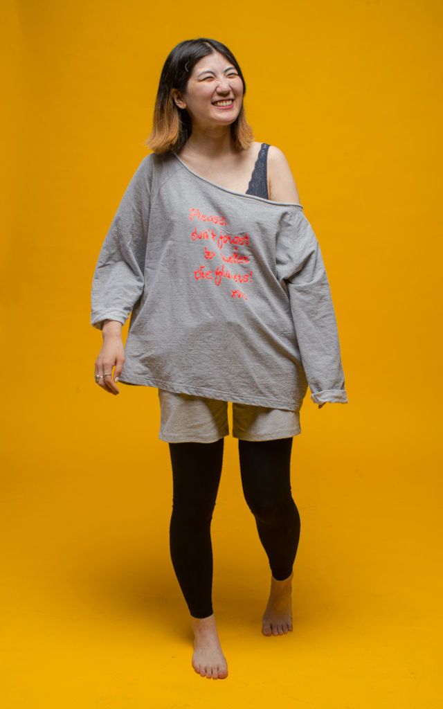A long, flowing, grey 5XL T-Shirt with a neon-orange print saying "Please don't forget to water the flowers, Xoxo!", worn by a female model in front of a yellow background.